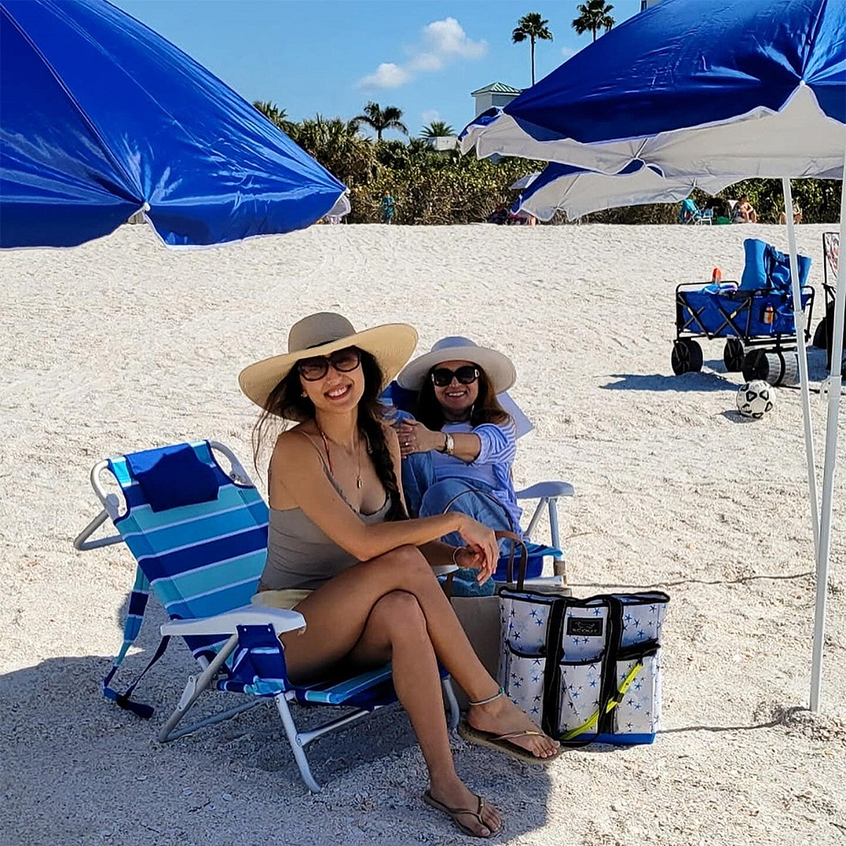 Two women smiling and relaxing on a sunny beach under blue umbrellas, enjoying a perfect day with sandy shores and clear skies.