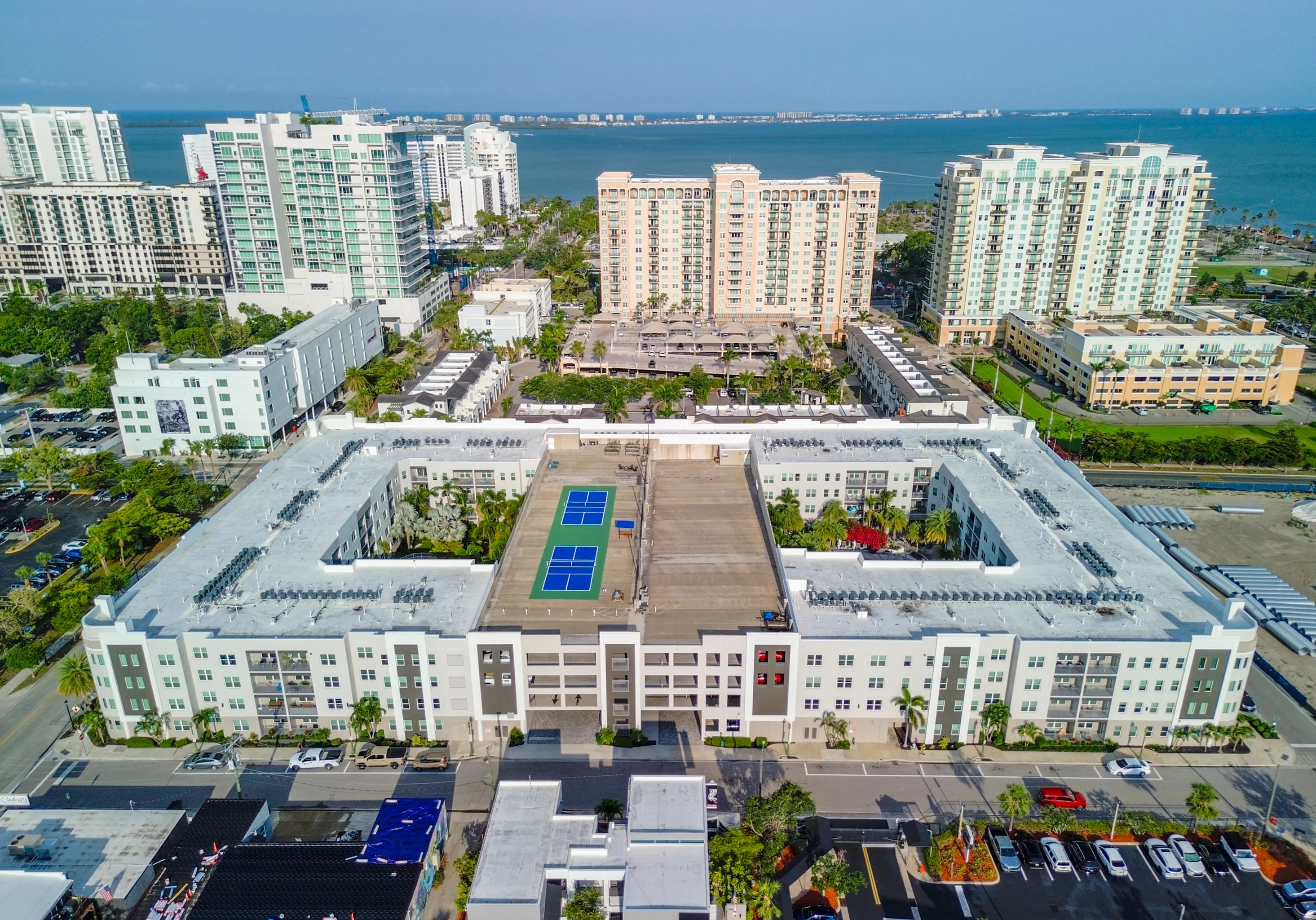 Aerial view of a contemporary apartment complex featuring multiple buildings, rooftop tennis courts, and stunning waterfront cityscape.