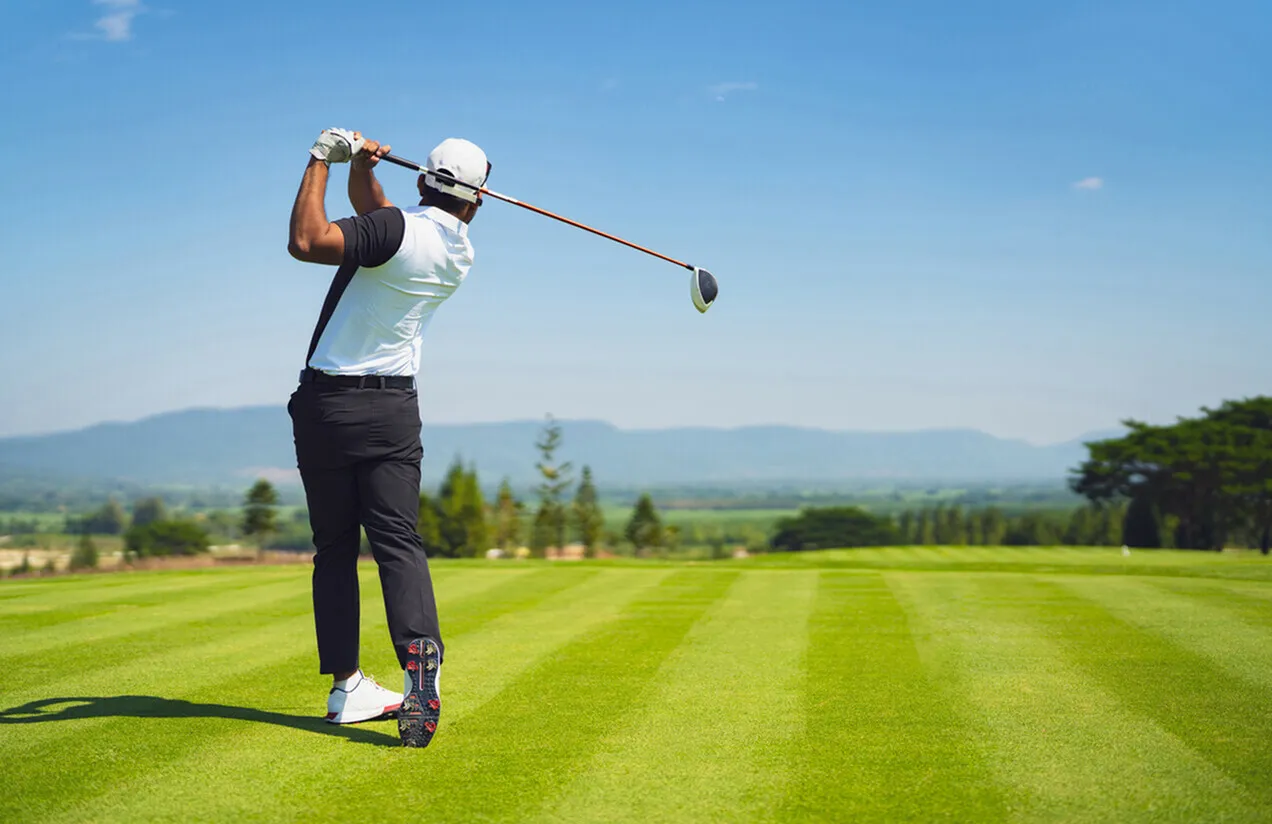 Vibrant golf course scenery with a player executing a perfect swing under clear blue skies, surrounded by lush greenery and distant mountains.