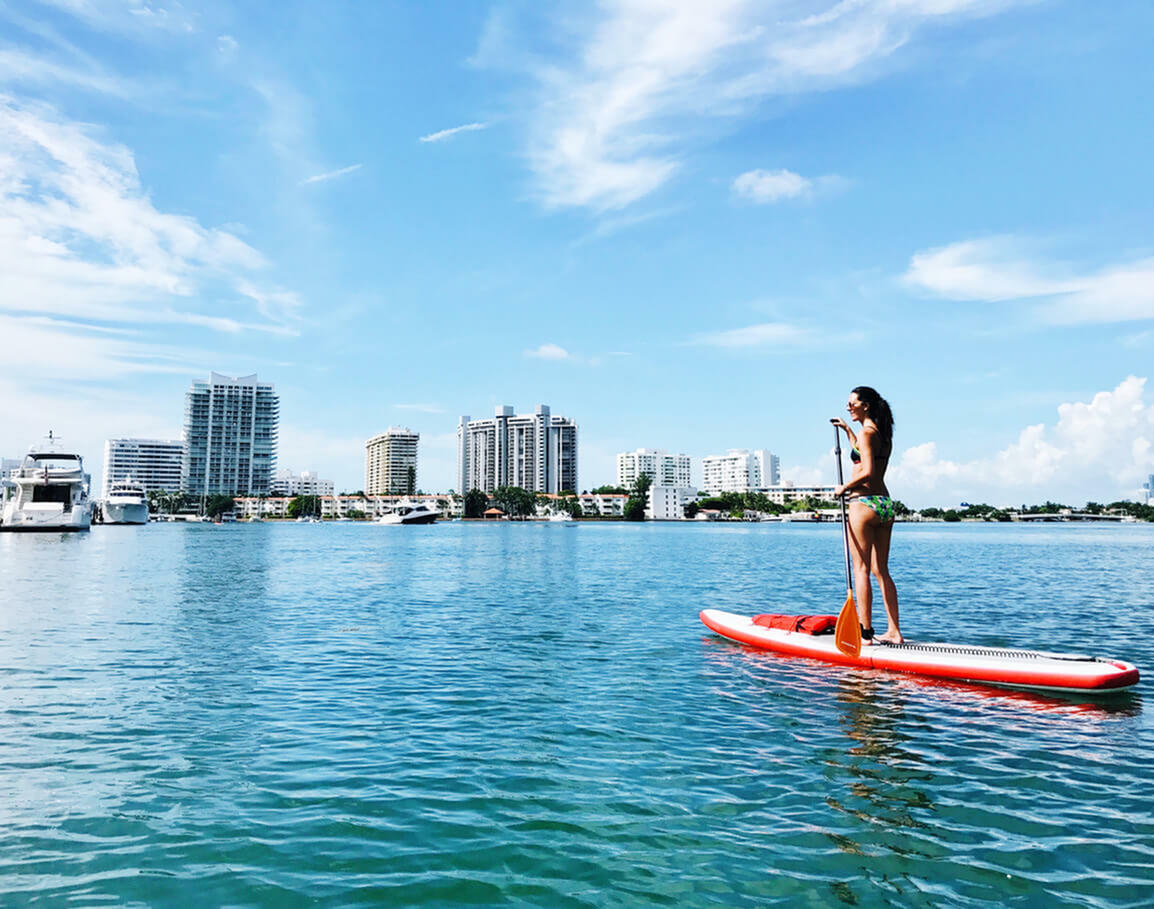 Woman paddleboarding on serene blue water with a stunning skyline of modern high-rise buildings, capturing the essence of waterfront urban living in luxury.