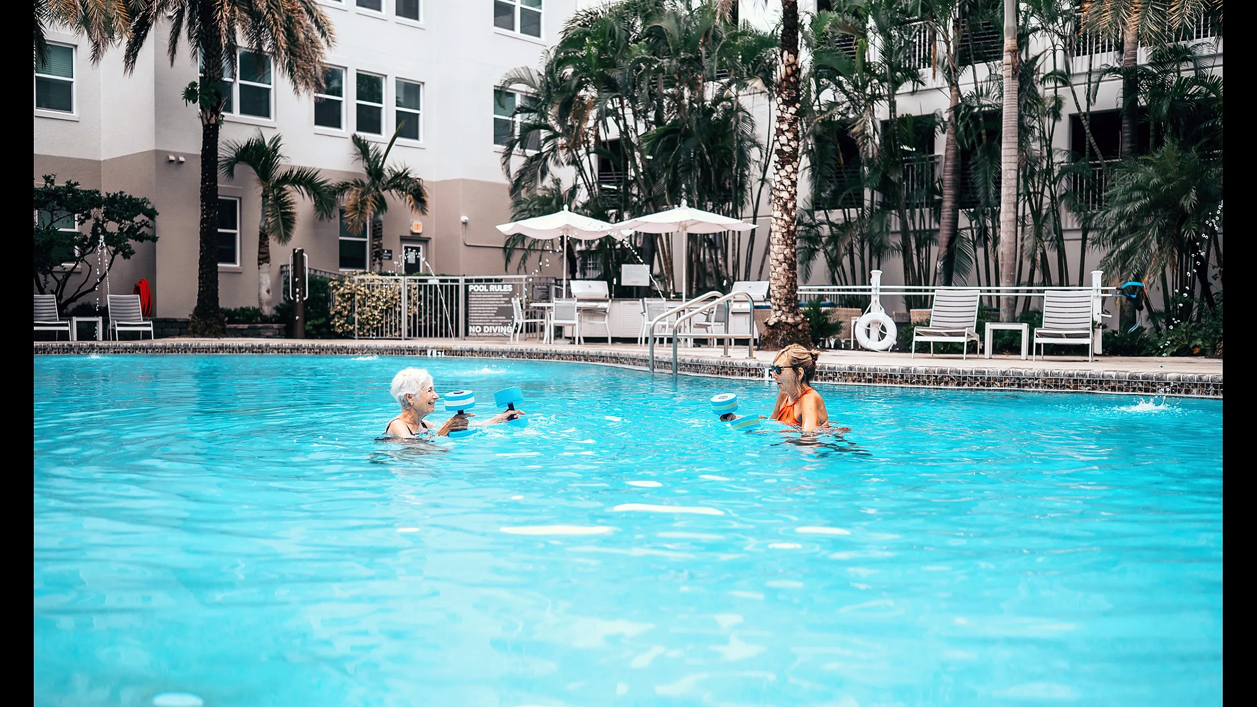 Inviting apartment pool area with vibrant blue water and lush palm trees, offering a relaxing oasis for residents enjoying aqua exercises.