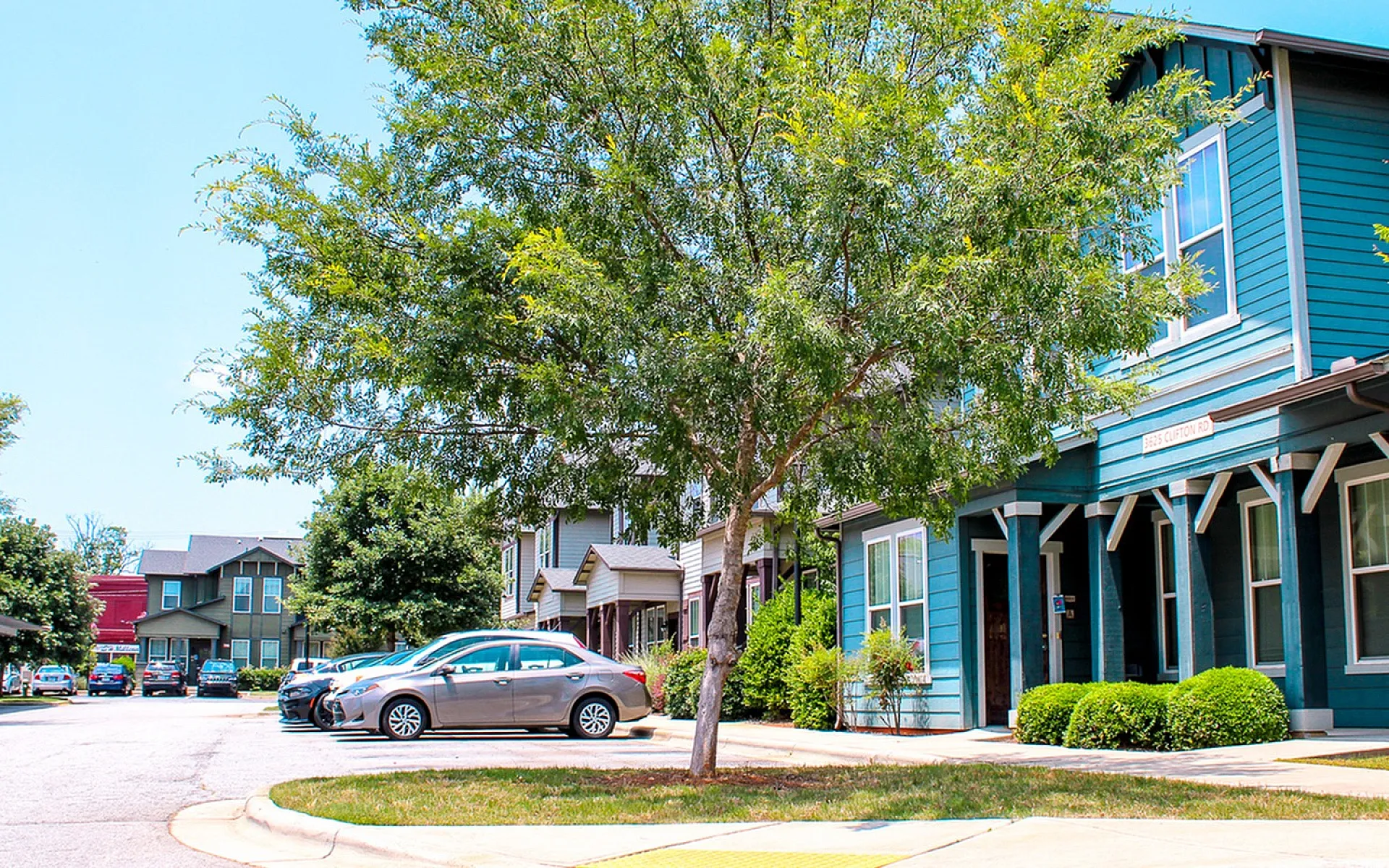 Charming suburban neighborhood with colorful townhouses, vibrant greenery, and ample parking space under a clear blue sky.
