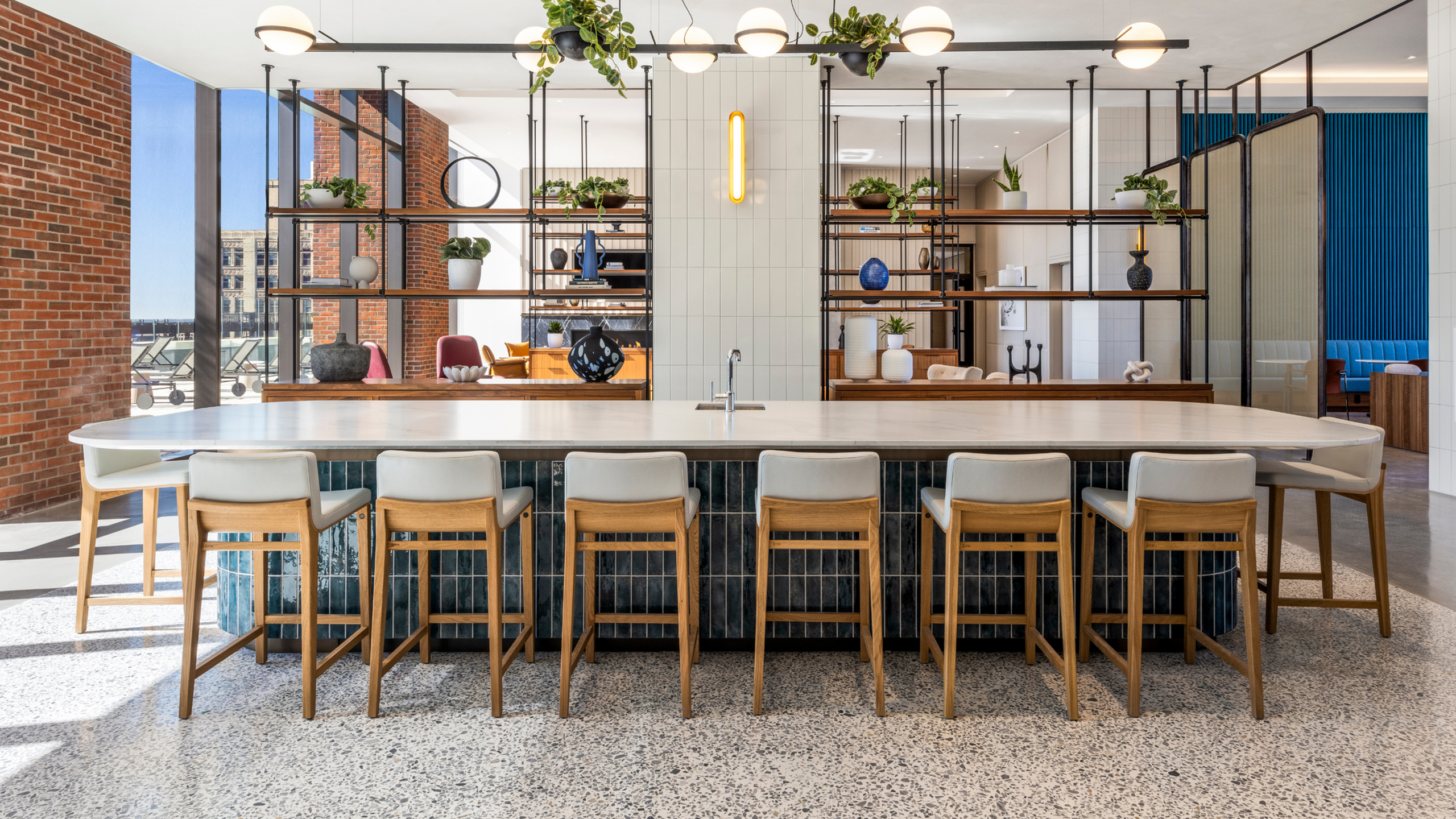 Chic and modern common area featuring a long marble countertop with high-backed stools, stylish shelving with plants, and warm natural light.