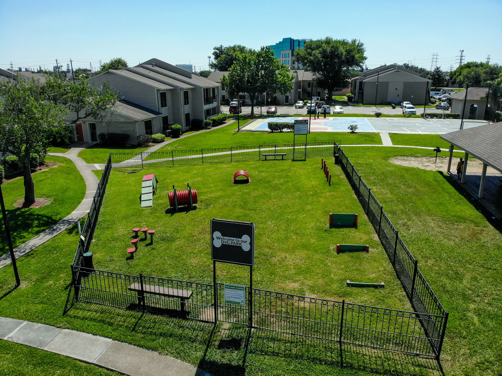 Vibrant apartment community showcasing a spacious, fenced dog park with agility equipment, surrounded by modern residential buildings and lush greenery.