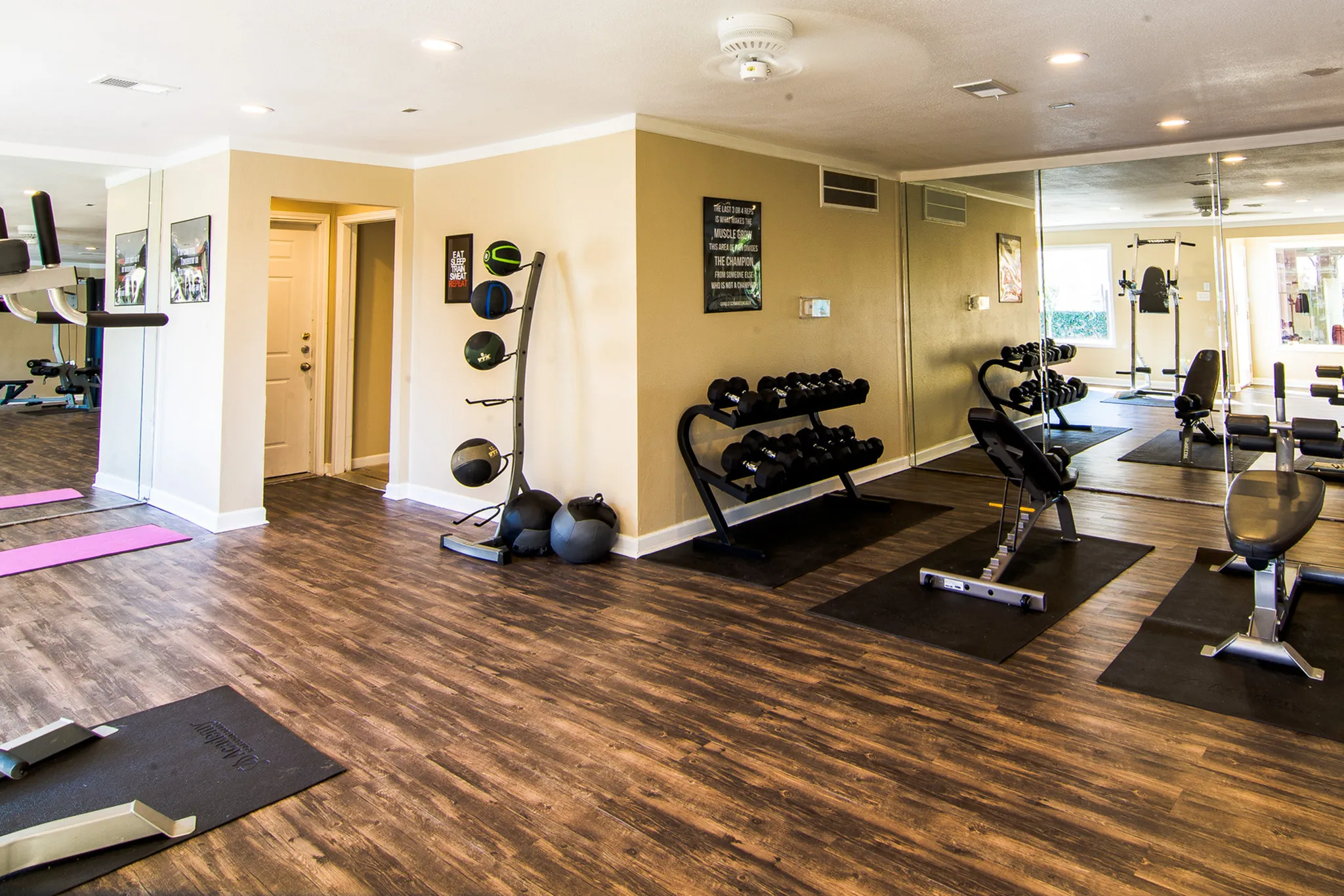 Bright and modern apartment gym with sleek wooden flooring, wall mirrors, and various exercise equipment including dumbbells, medicine balls, and workout benches.