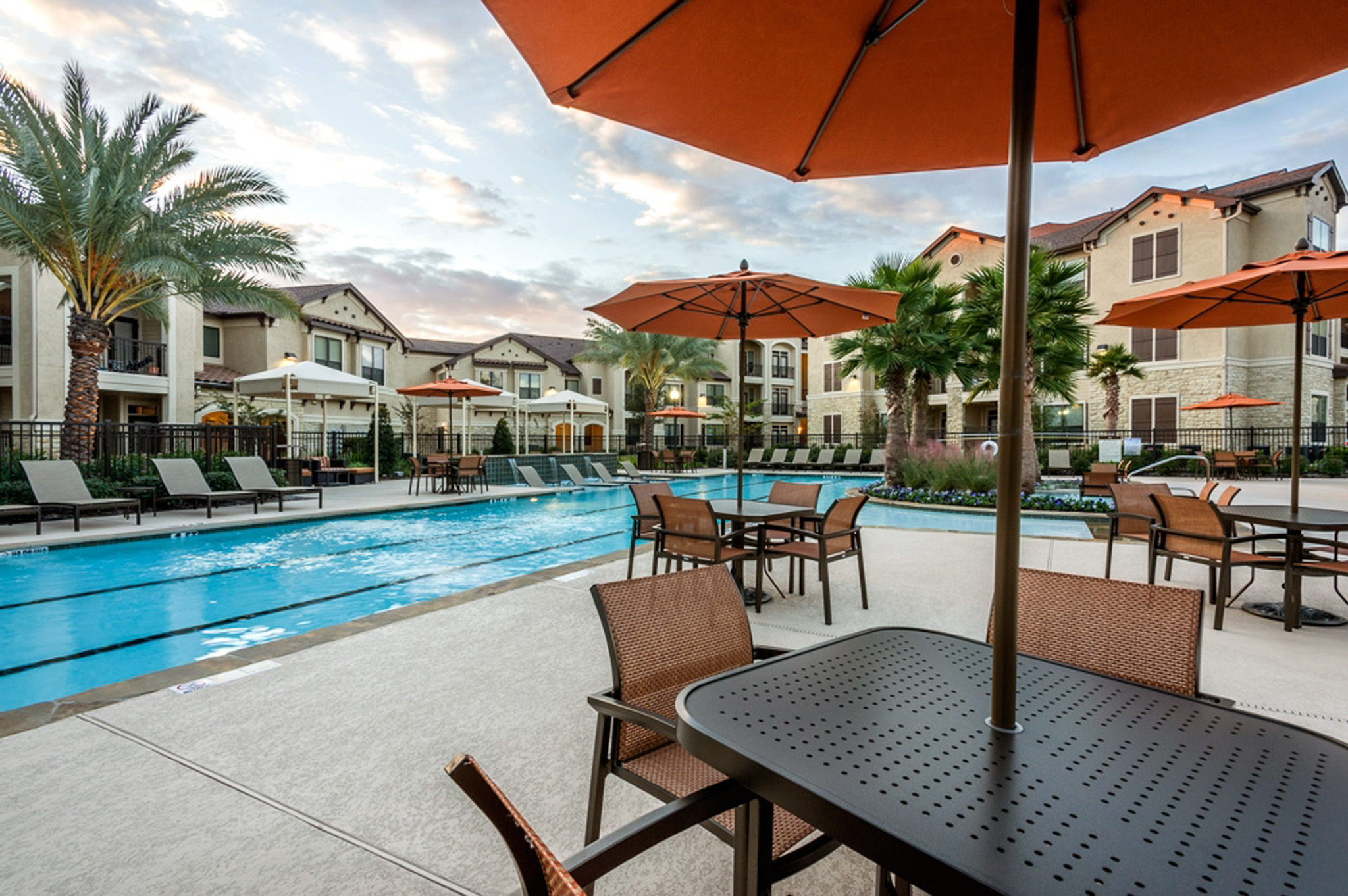 Elegant apartment complex with a serene poolside setting, featuring sun loungers, vibrant umbrellas, and lush palm trees.