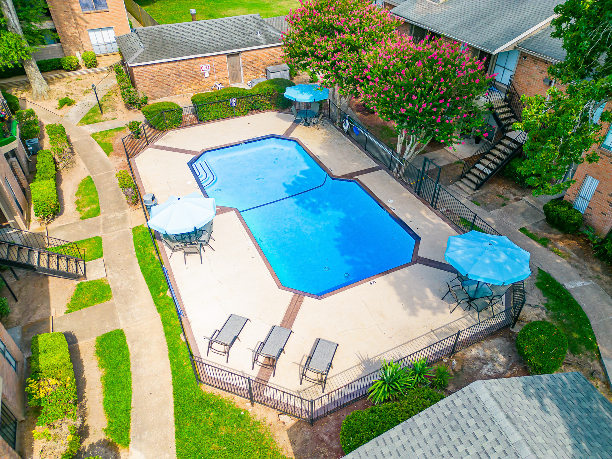Charming apartment complex with a sparkling blue pool, surrounded by lush greenery and vibrant flowers, offers a serene outdoor oasis with stylish lounge chairs and shaded seating areas.