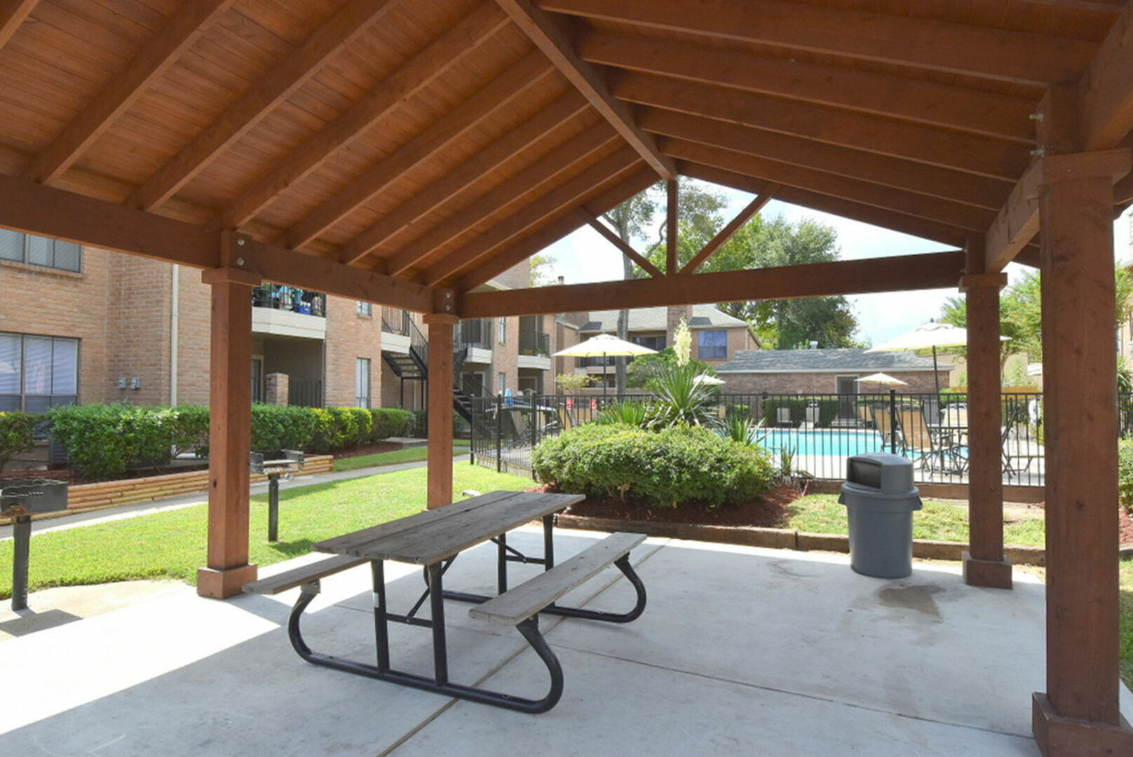 Charming outdoor pavilion with wooden beams, picnic table, and scenic view of lush gardens and a sparkling pool.