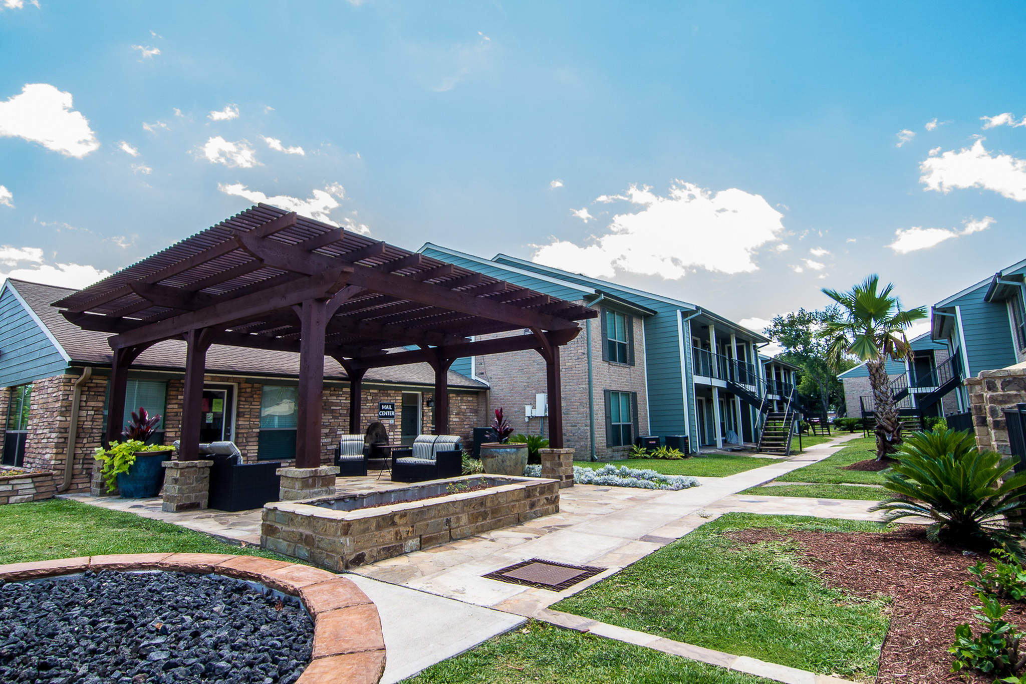 Inviting apartment community with lush landscaping, outdoor seating area, and modern architecture under a bright blue sky.