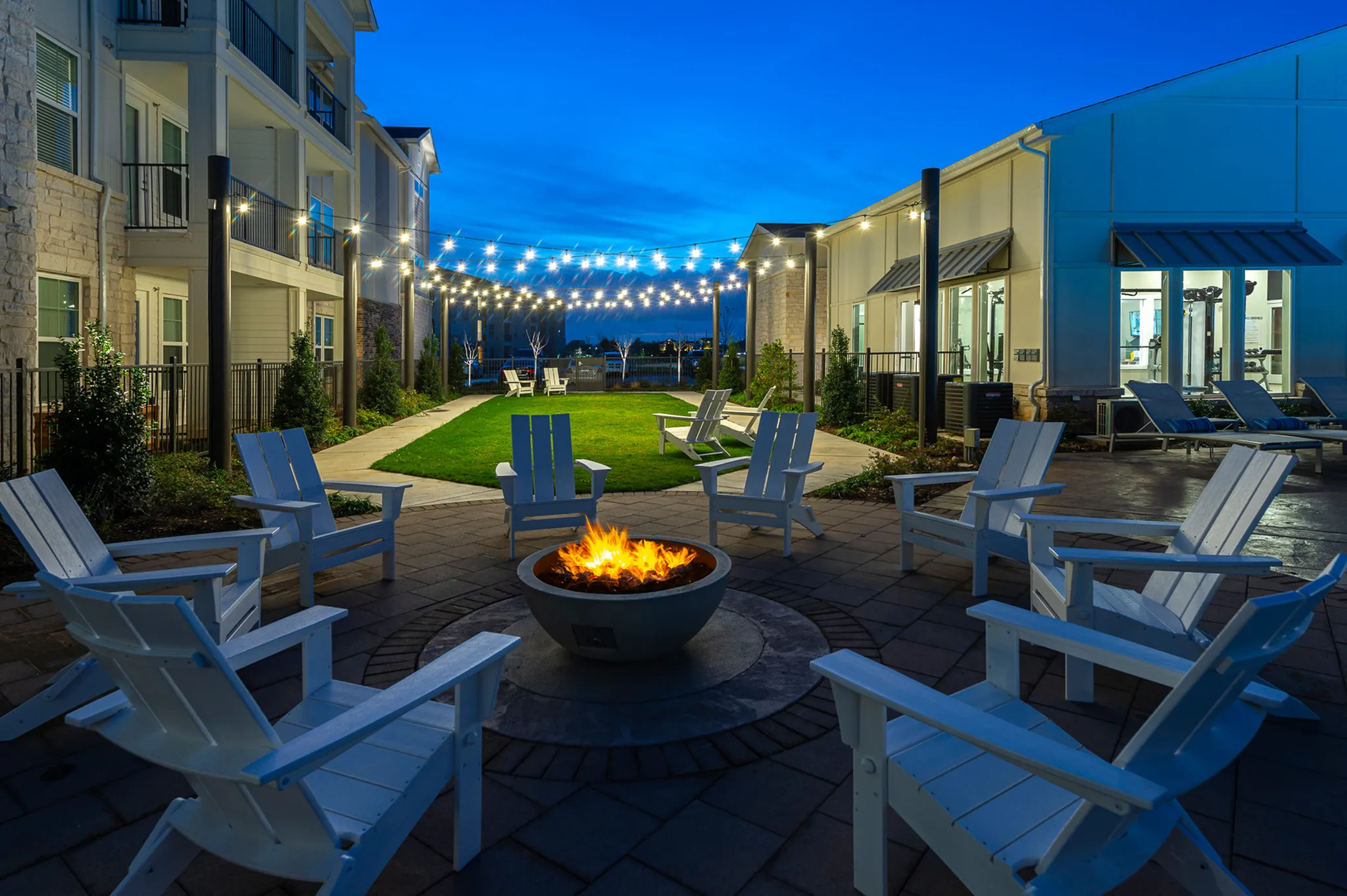 Inviting outdoor space with a cozy fire pit surrounded by white chairs, under twinkling string lights, perfect for evening relaxation.