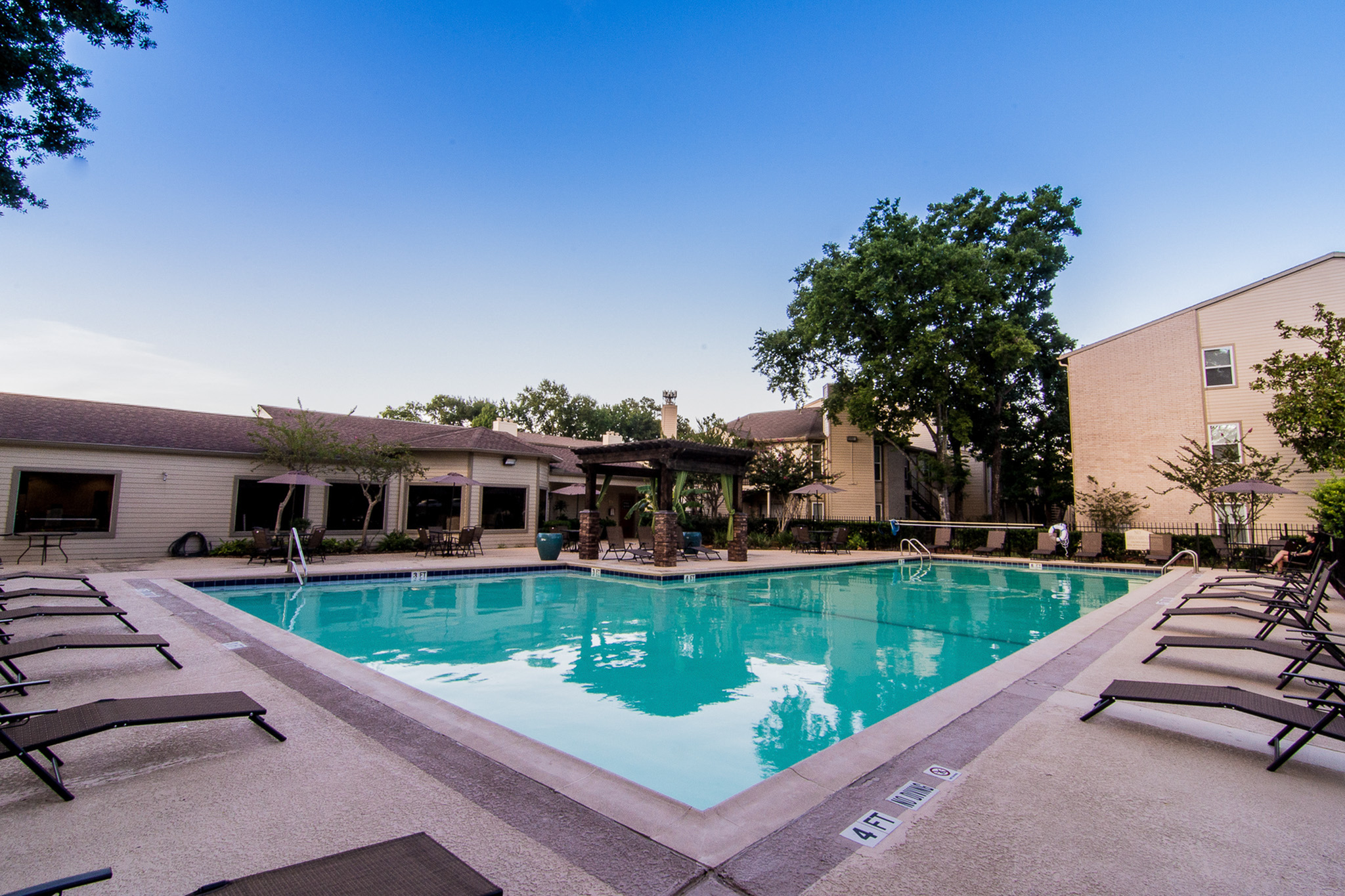 Inviting outdoor pool area surrounded by lounge chairs, with a charming pergola and modern apartment buildings under a clear blue sky.