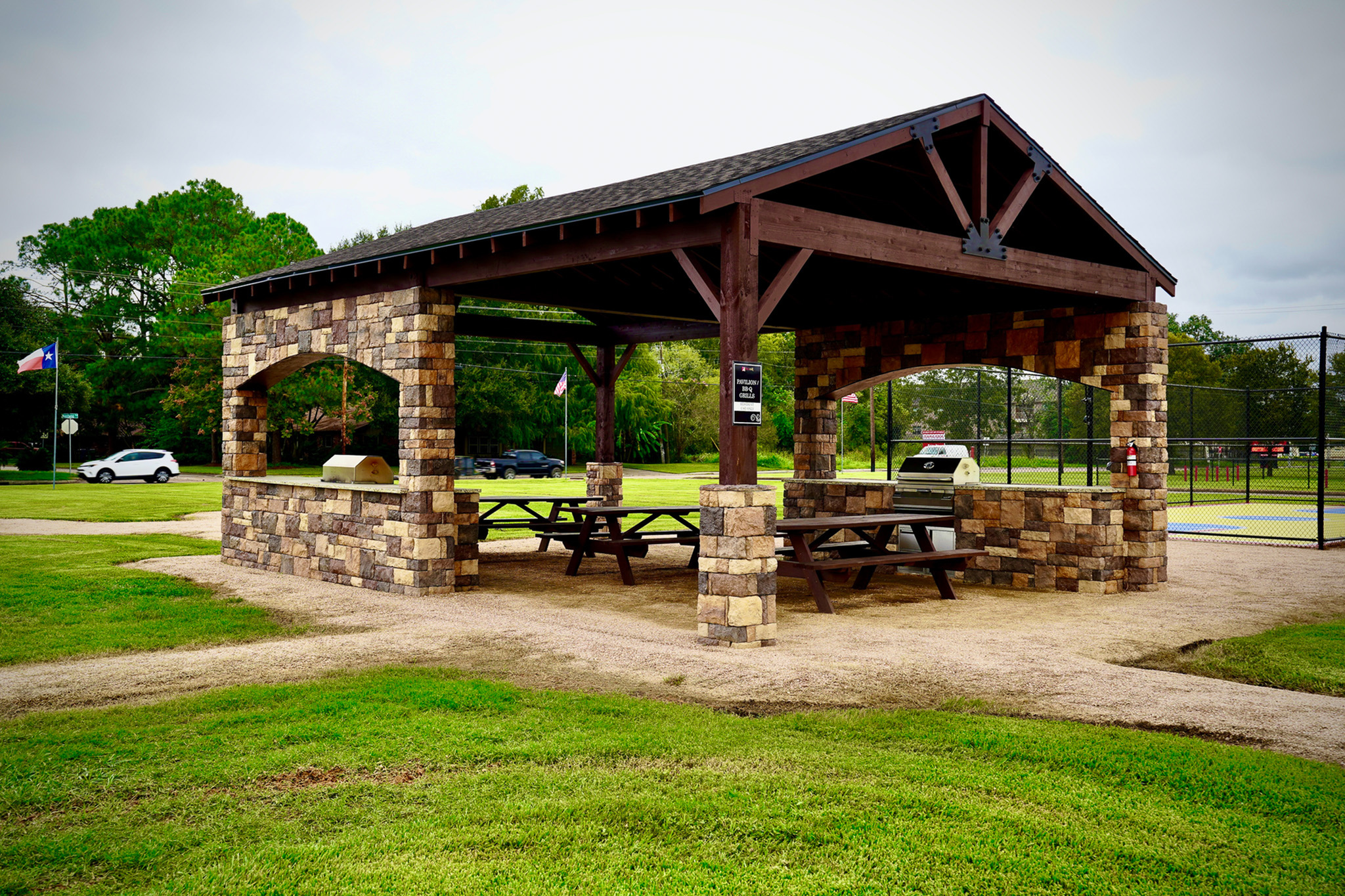 Charming outdoor pavilion with rustic stone design, featuring picnic tables and a modern grill, set in a lush green park.