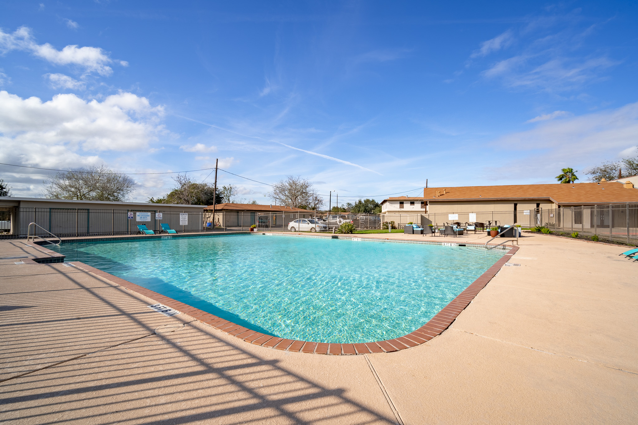 Inviting apartment community pool area with crystal-clear water, surrounded by a spacious deck and lounge chairs under a clear blue sky.