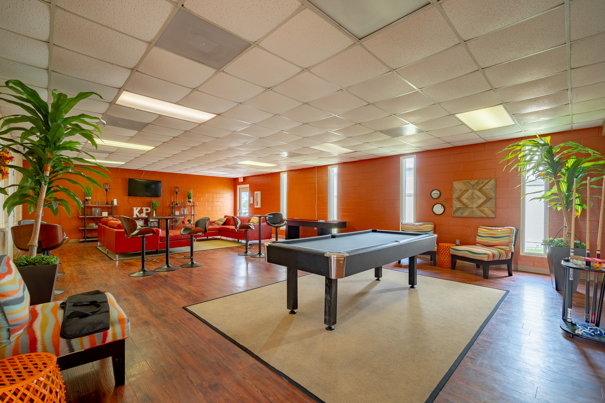 Vibrant game room featuring a sleek pool table, stylish seating, and lush green plants, with warm orange walls creating an inviting atmosphere.