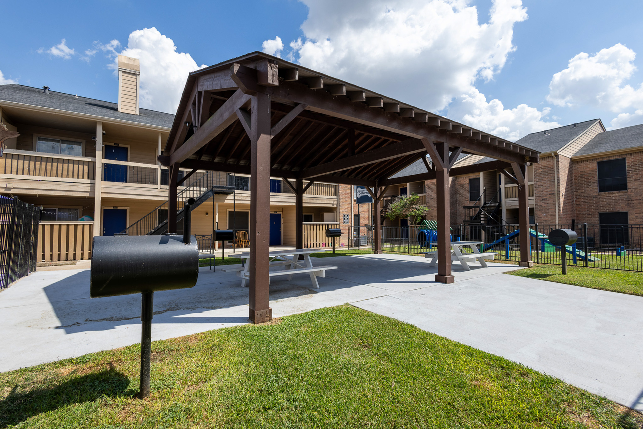 Charming apartment complex with a shaded wooden pergola, picnic tables, and barbecue grills, perfect for outdoor gatherings.