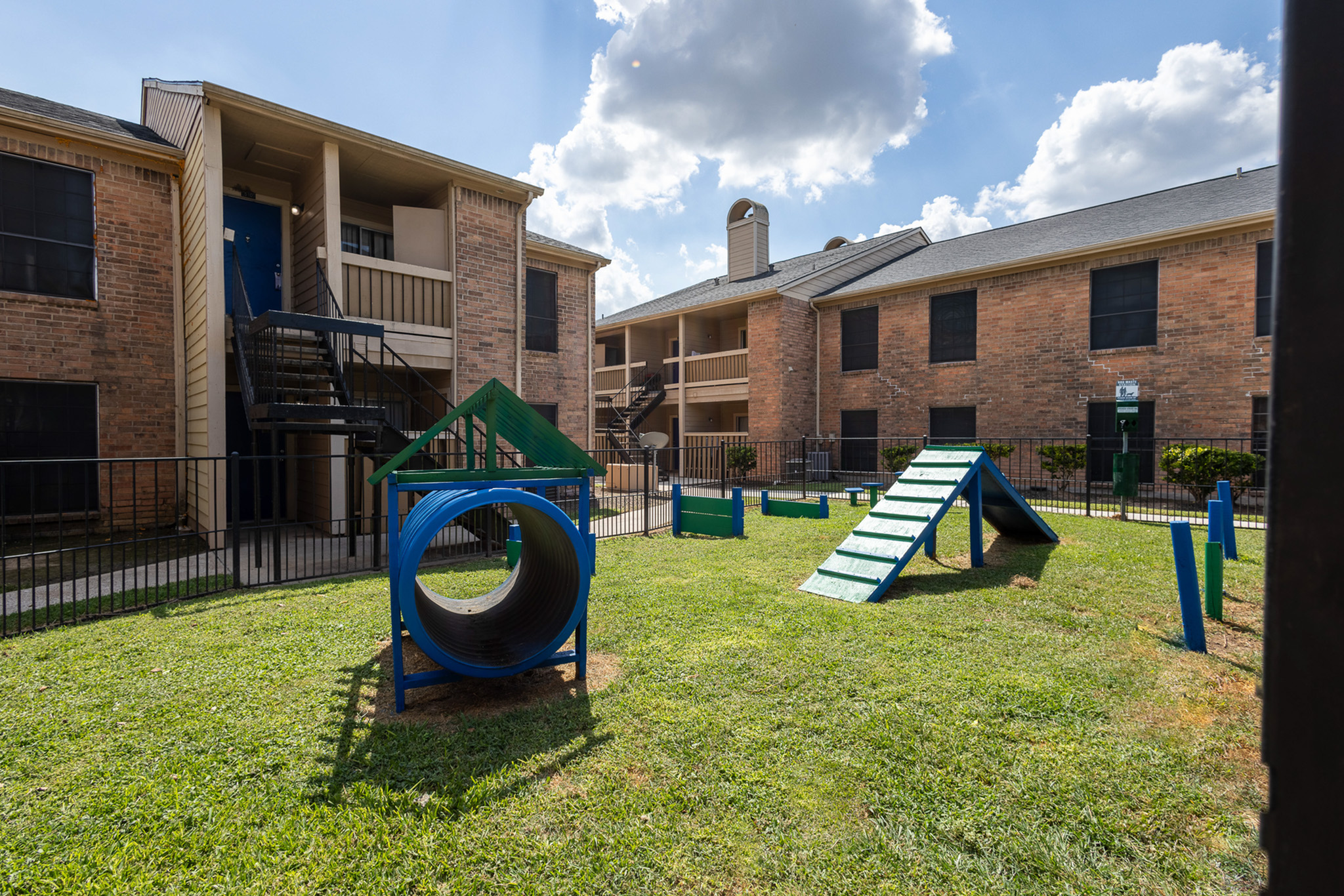Inviting apartment exterior with a dog-friendly play area, complete with agility obstacles and lush green grass under a bright blue sky.