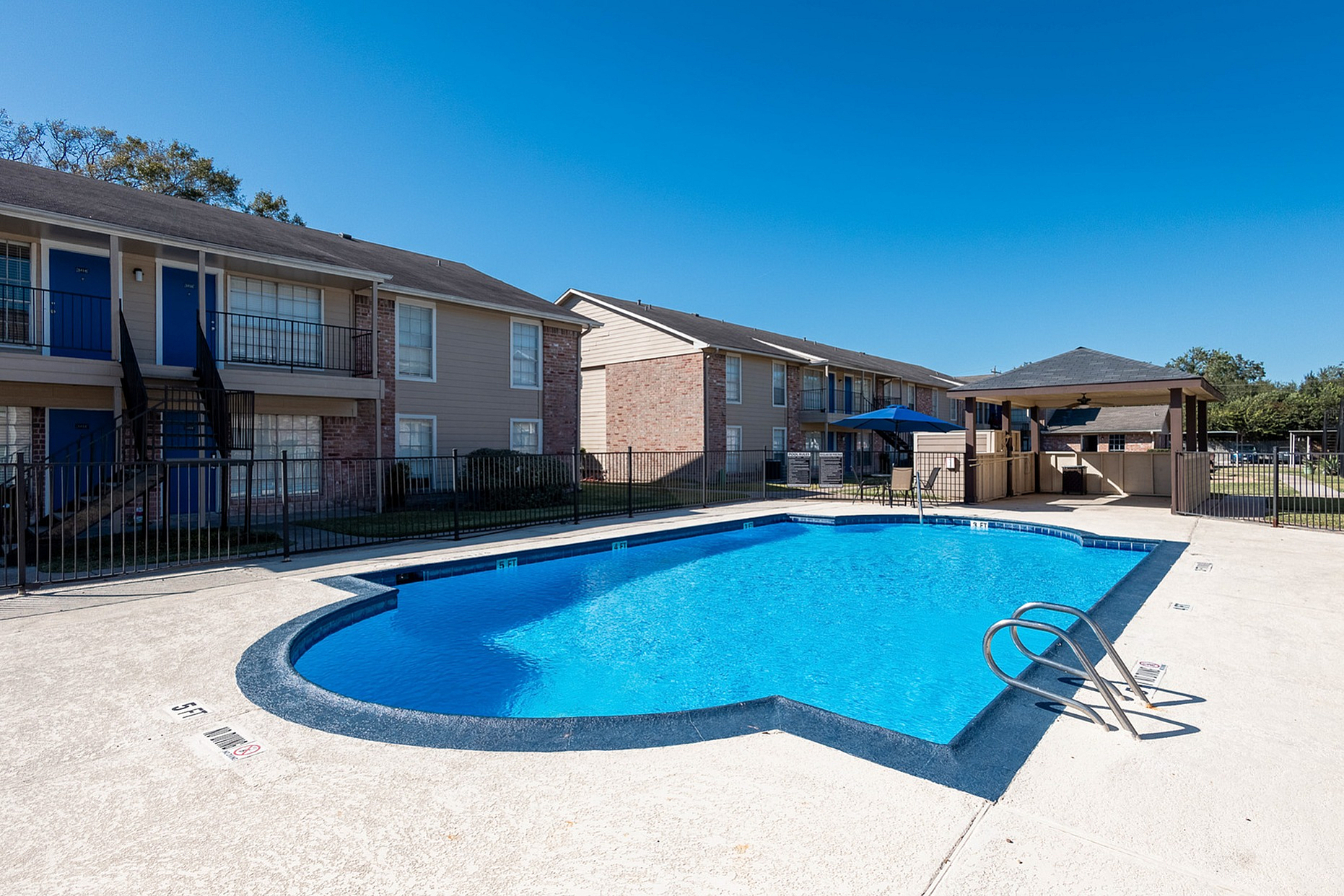 Inviting apartment complex with a sparkling blue pool, surrounded by modern brick buildings and sunny skies.