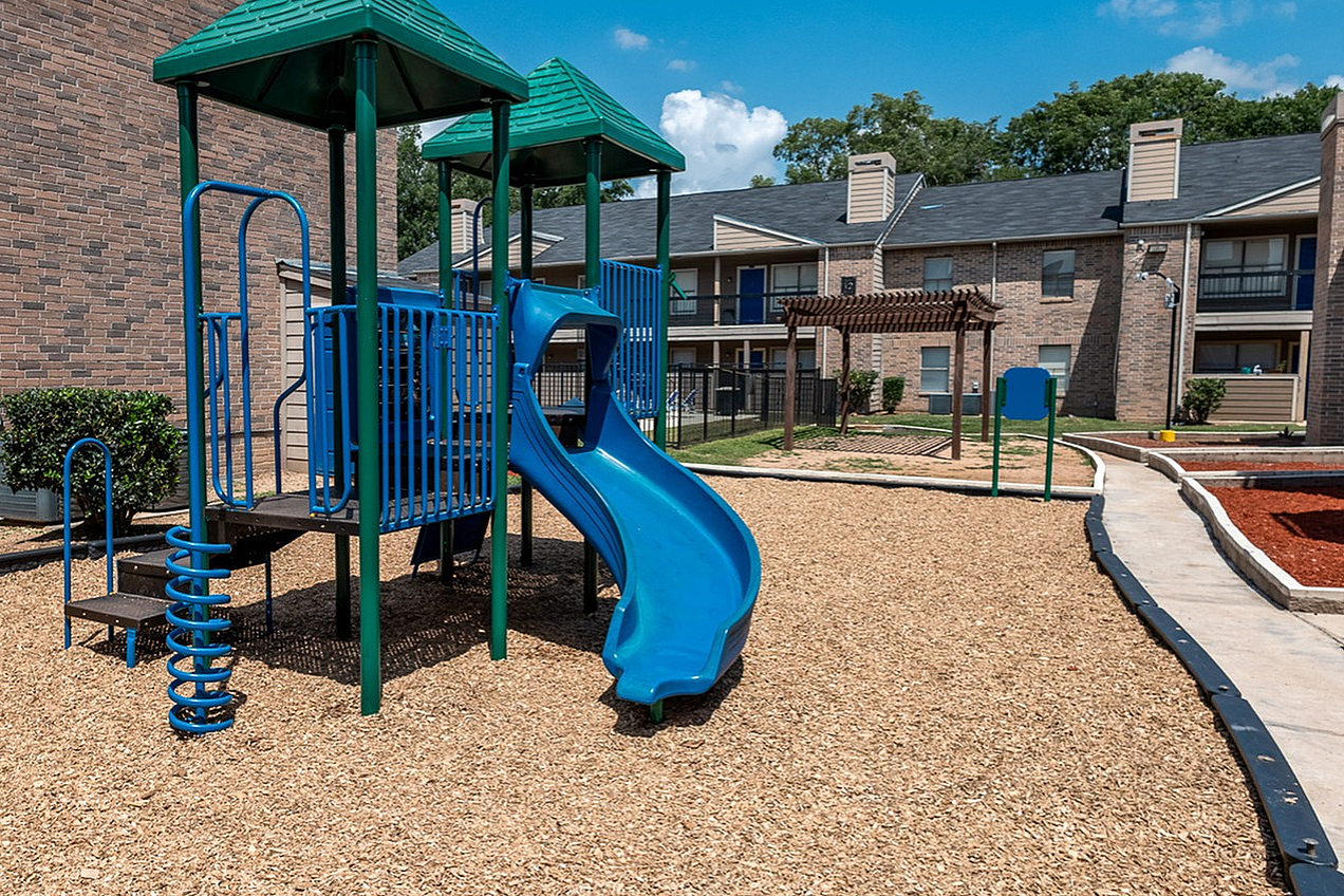 Inviting apartment complex with a vibrant playground featuring a colorful slide, surrounded by lush greenery and modern brick buildings.