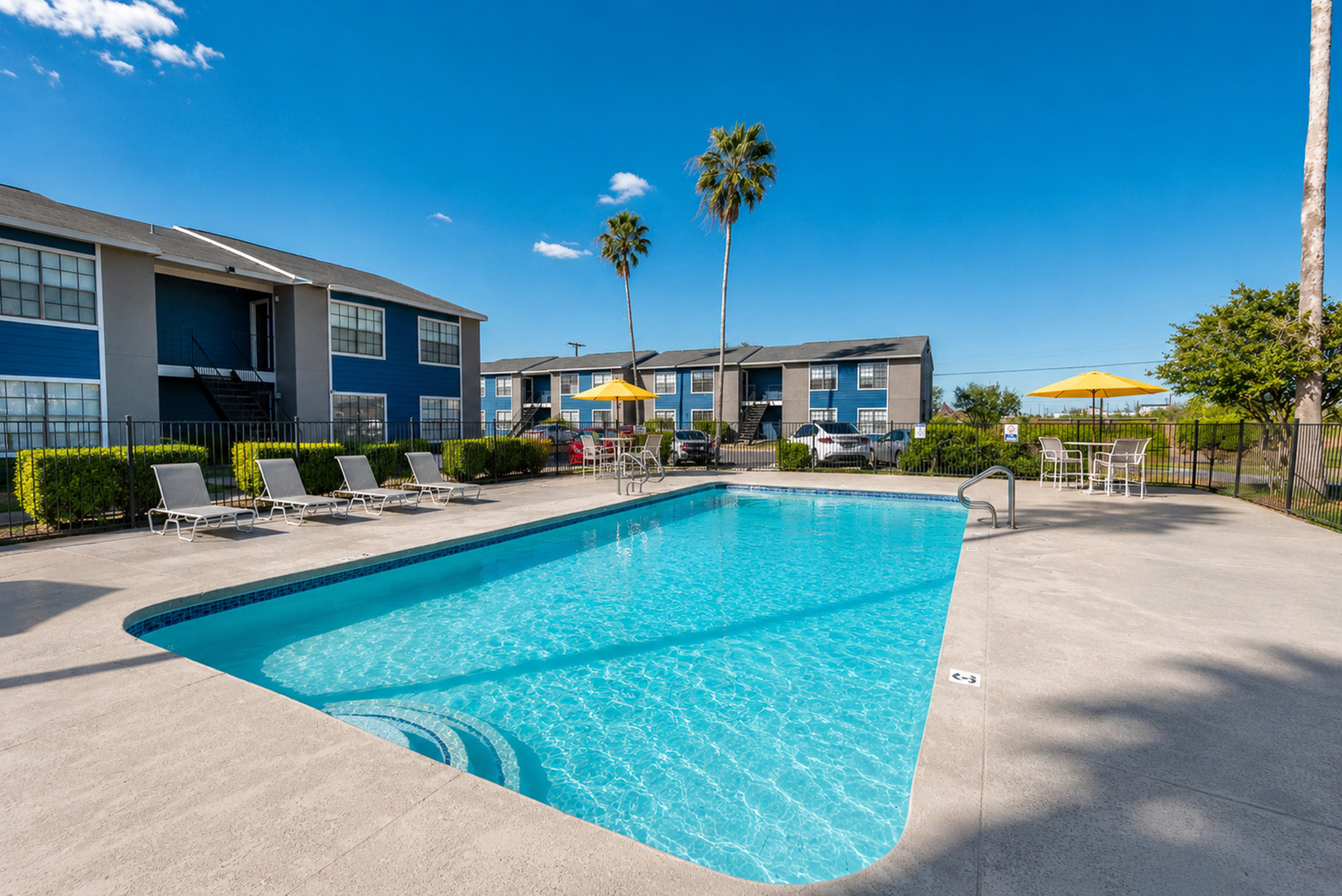 Inviting apartment complex with a sparkling outdoor pool, surrounded by lush greenery and comfortable lounge chairs under a clear blue sky.