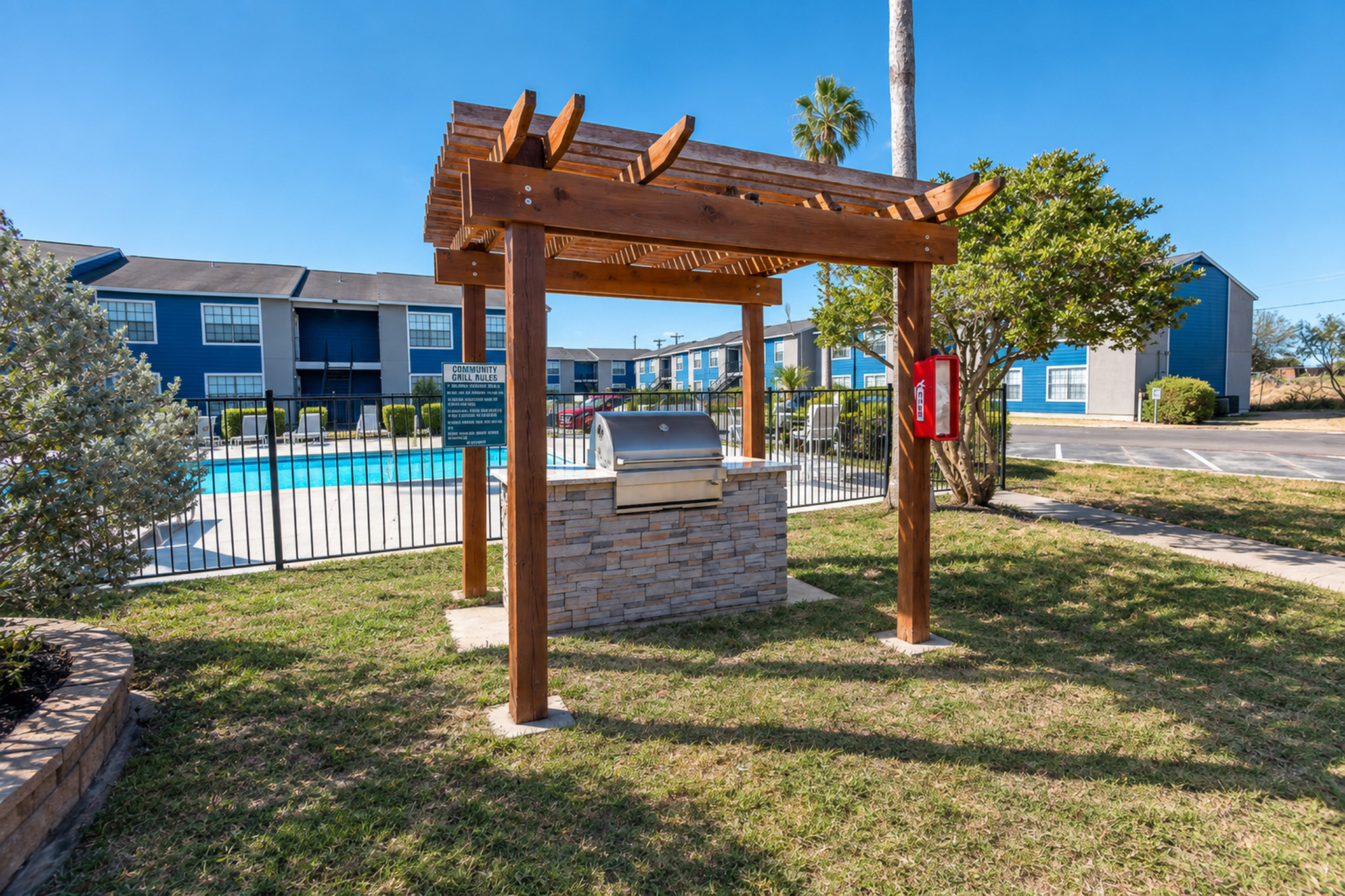 Inviting outdoor barbecue area with a modern grill, lush greenery, and a sparkling blue pool in a vibrant community setting.