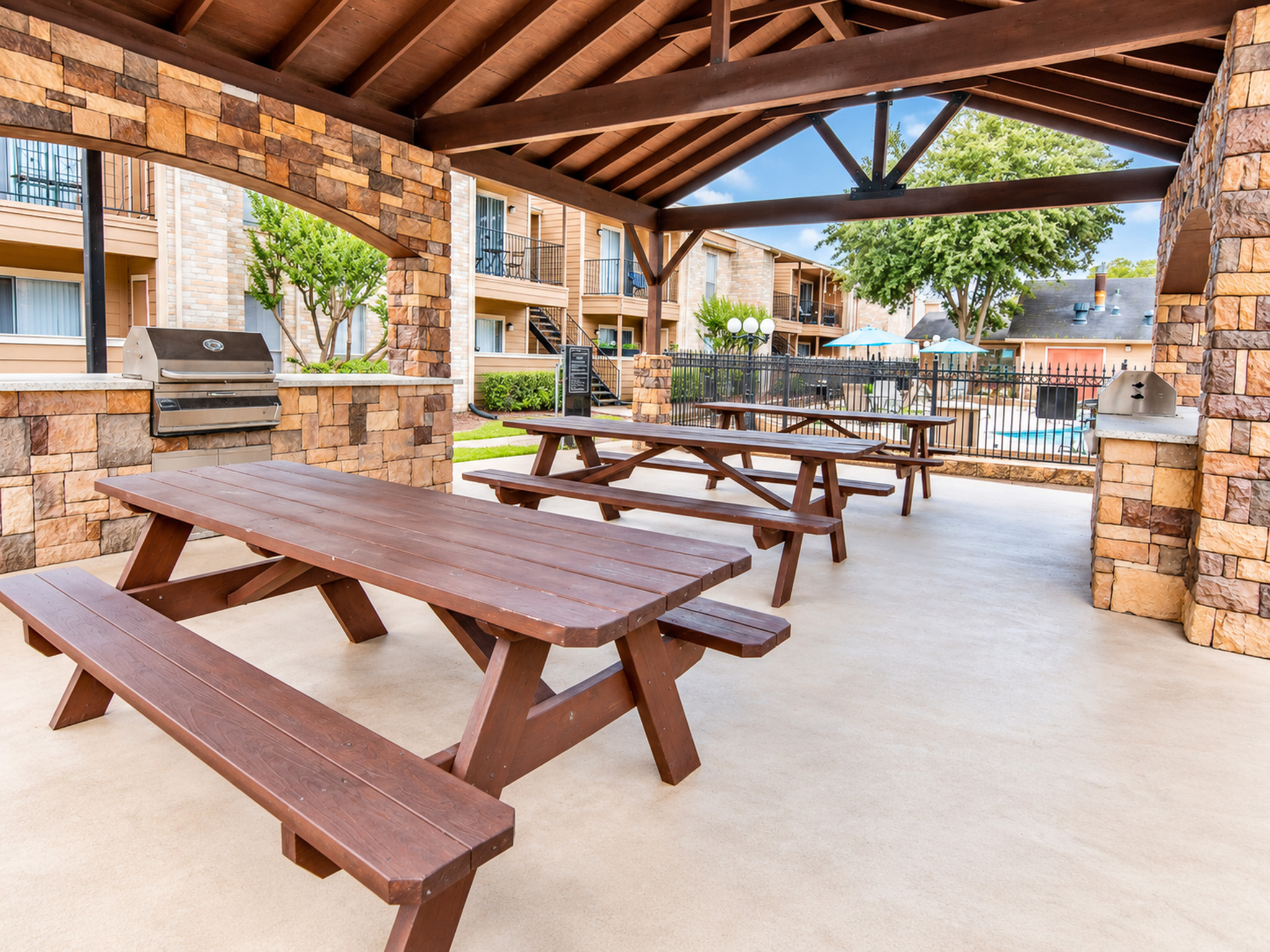 Charming outdoor pavilion with wooden picnic tables and a stone barbecue area, ideal for gatherings and overlooking a beautiful pool.