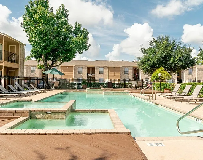 Inviting pool area with sun loungers in a serene apartment complex, surrounded by lush greenery and clear blue skies.
