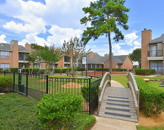 Charming apartment complex with lush green landscaping, a quaint bridge, and red-brick buildings under a vibrant blue sky.