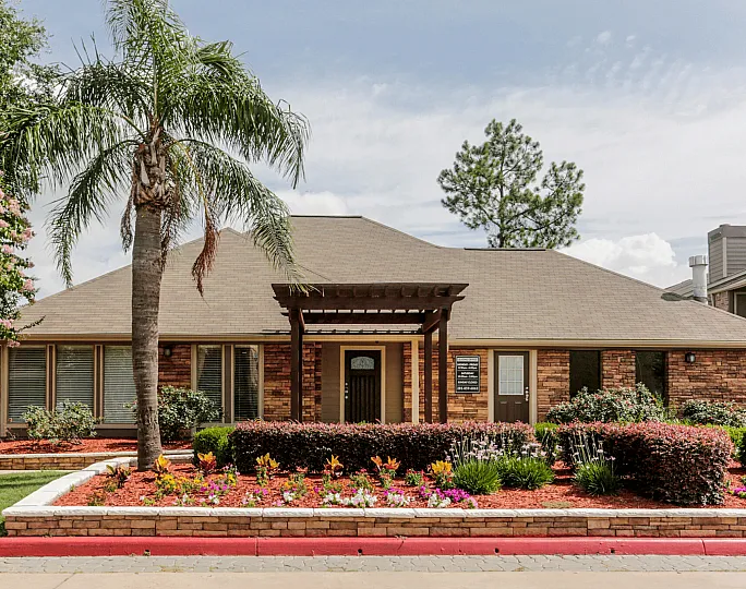 Charming brick apartment building with lush landscaping and vibrant flower beds, highlighted by a welcoming palm tree entrance.