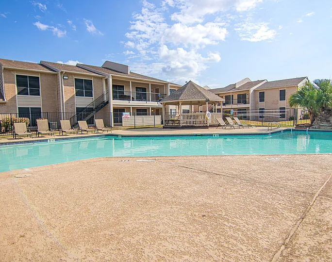 Inviting apartment complex with a sparkling swimming pool, surrounded by lounge chairs and a charming gazebo, under a bright blue sky.