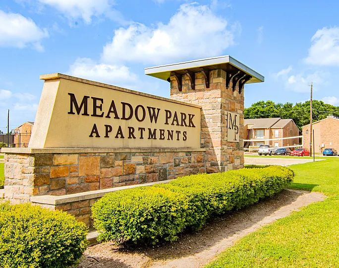 Charming Meadow Park Apartments entry sign with beautifully landscaped surroundings and residential buildings in the background, under a bright blue sky.