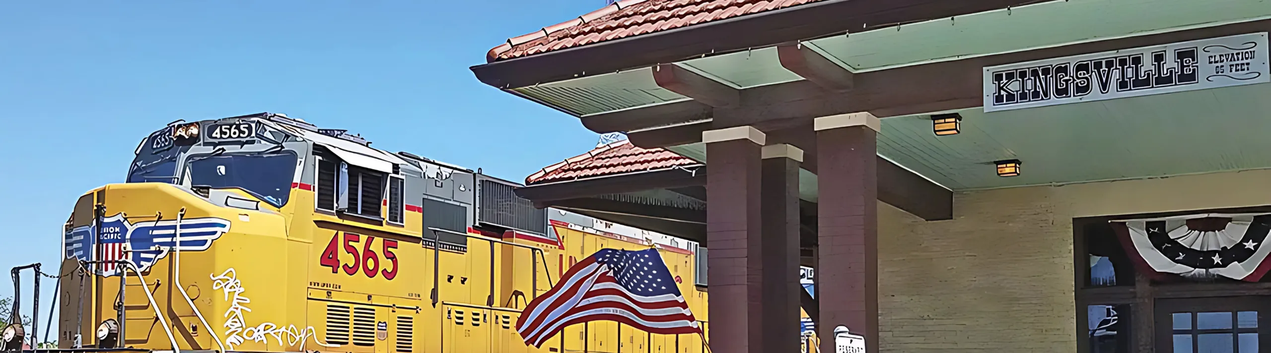 Vibrant urban scene with a yellow Union Pacific train passing by the historic Kingsville station under a clear blue sky, adorned with American flag decorations.