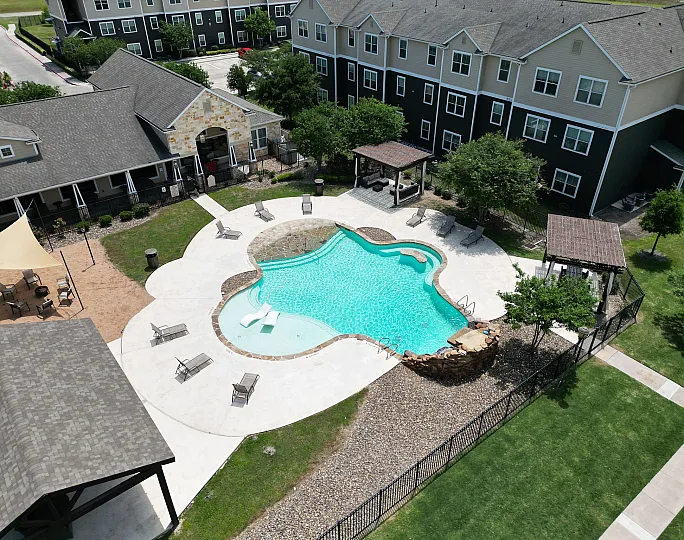 Aerial view of a luxurious apartment complex featuring a sparkling kidney-shaped pool, surrounded by lounge chairs and shaded seating areas, offering a resort-style living experience.
