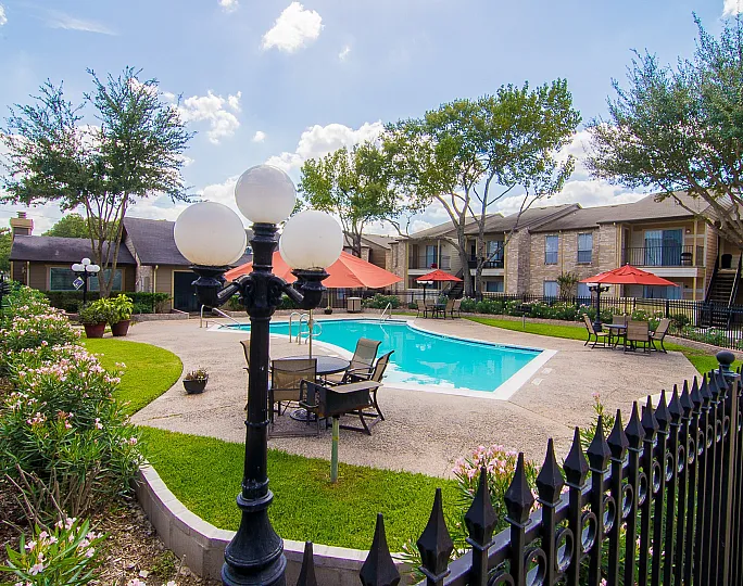Charming apartment complex with a sparkling pool, cozy patio seating, and lush landscaping under a bright blue sky.