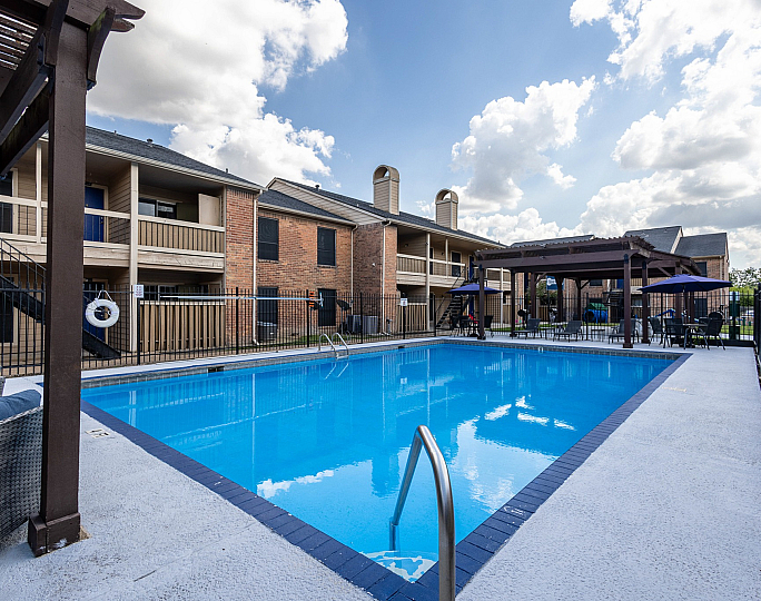 Modern apartment complex showcasing a sunlit swimming pool with stylish loungers and shaded seating areas, perfect for relaxation.