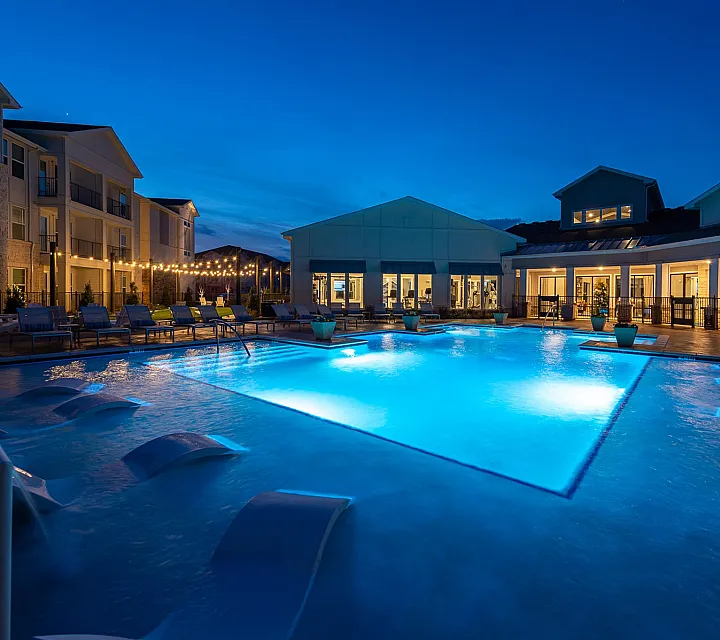 Luxurious apartment complex featuring a stunning illuminated pool area with comfortable lounge chairs, perfect for evening relaxation.