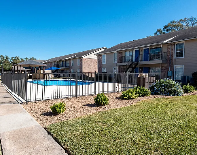 Charming apartment complex with a central swimming pool, surrounded by well-maintained lawns and beautiful landscaping under a clear blue sky.