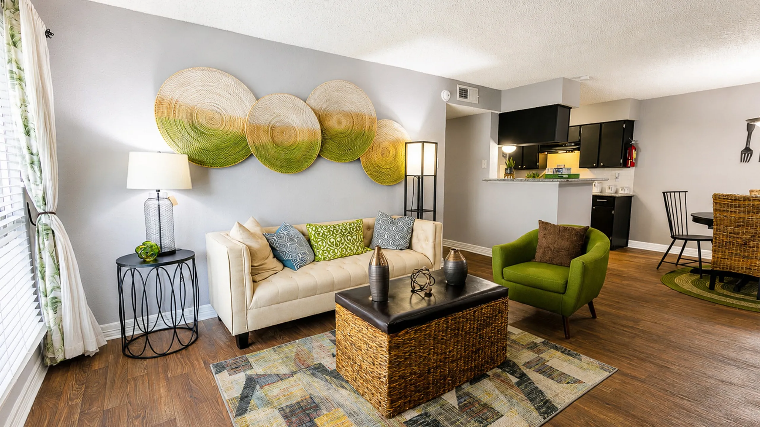 Chic living room with modern decor, featuring a cozy cream sofa, vibrant green chair, and stylish woven wall art, seamlessly connected to a sleek black kitchen.