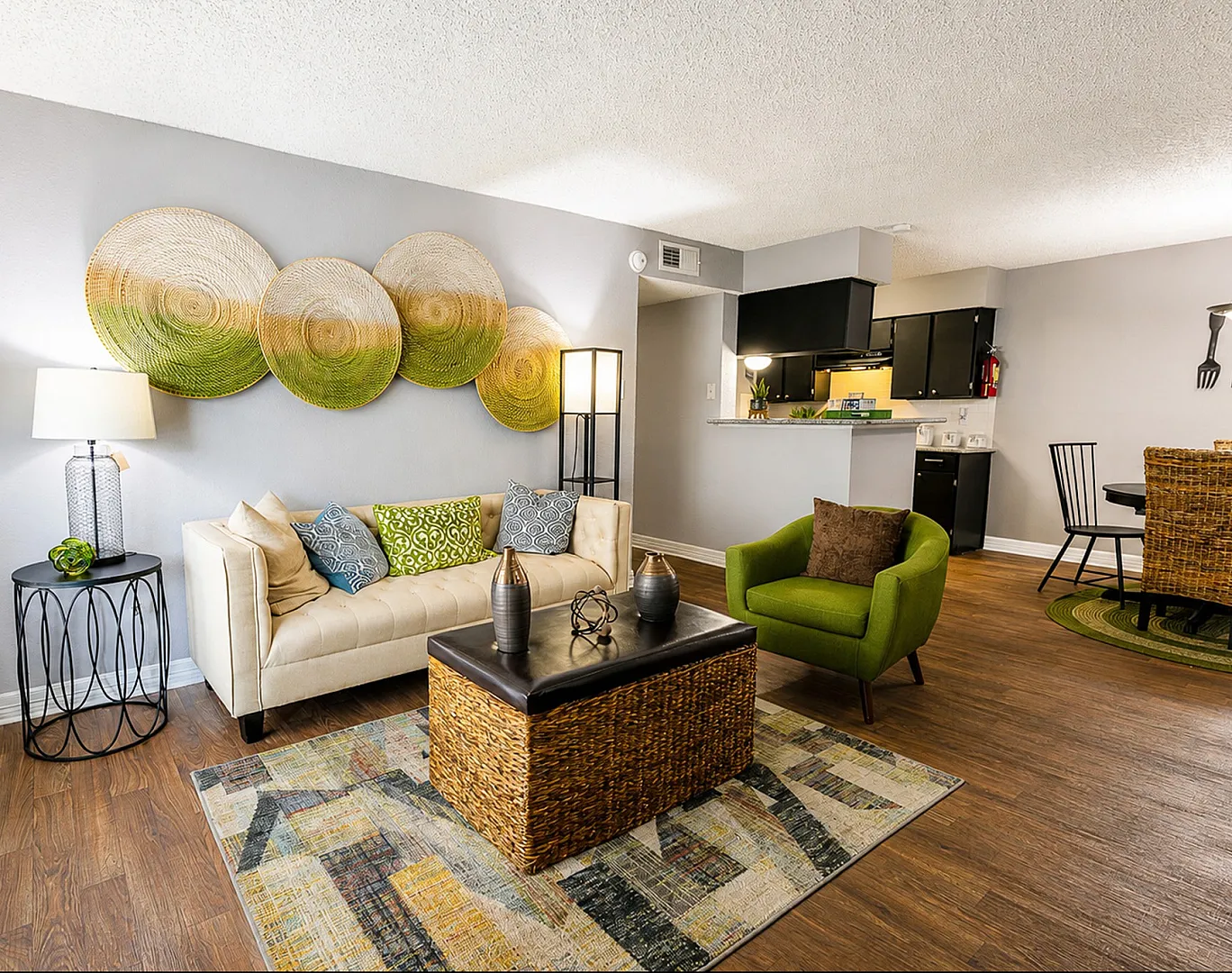 Inviting living space with modern decor, featuring a cozy beige sofa, stylish green armchair, and vibrant woven wall art. Open kitchen with sleek black cabinetry complements the warm wooden flooring.