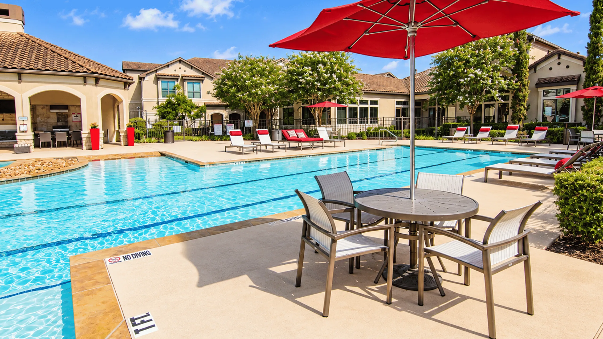 Pool and patio seating at Valencia Place apartments in Houston, TX with red umbrellas, lounge chairs and landscaped surroundings.