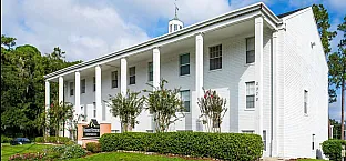 Elegant white colonial-style apartment building with grand columns and lush green landscaping under a bright sky.