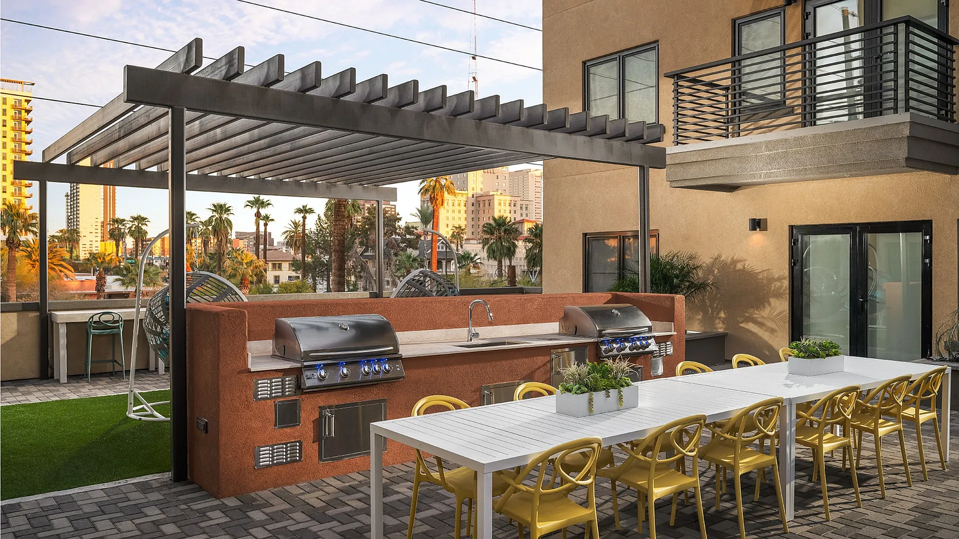 Chic outdoor patio with modern BBQ grills, a long dining table with vibrant yellow chairs, set against a backdrop of palm trees and a cityscape view.