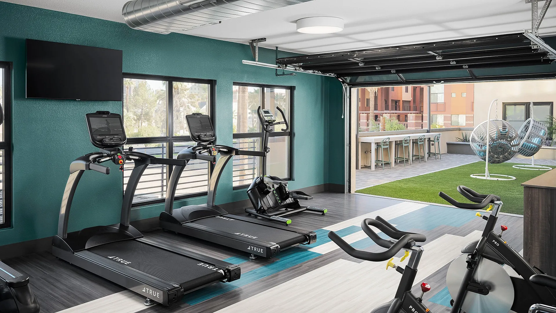 Modern fitness center with state-of-the-art treadmills and exercise bikes, overlooking a stylish outdoor lounge area with chic hanging chairs and a vibrant green lawn.