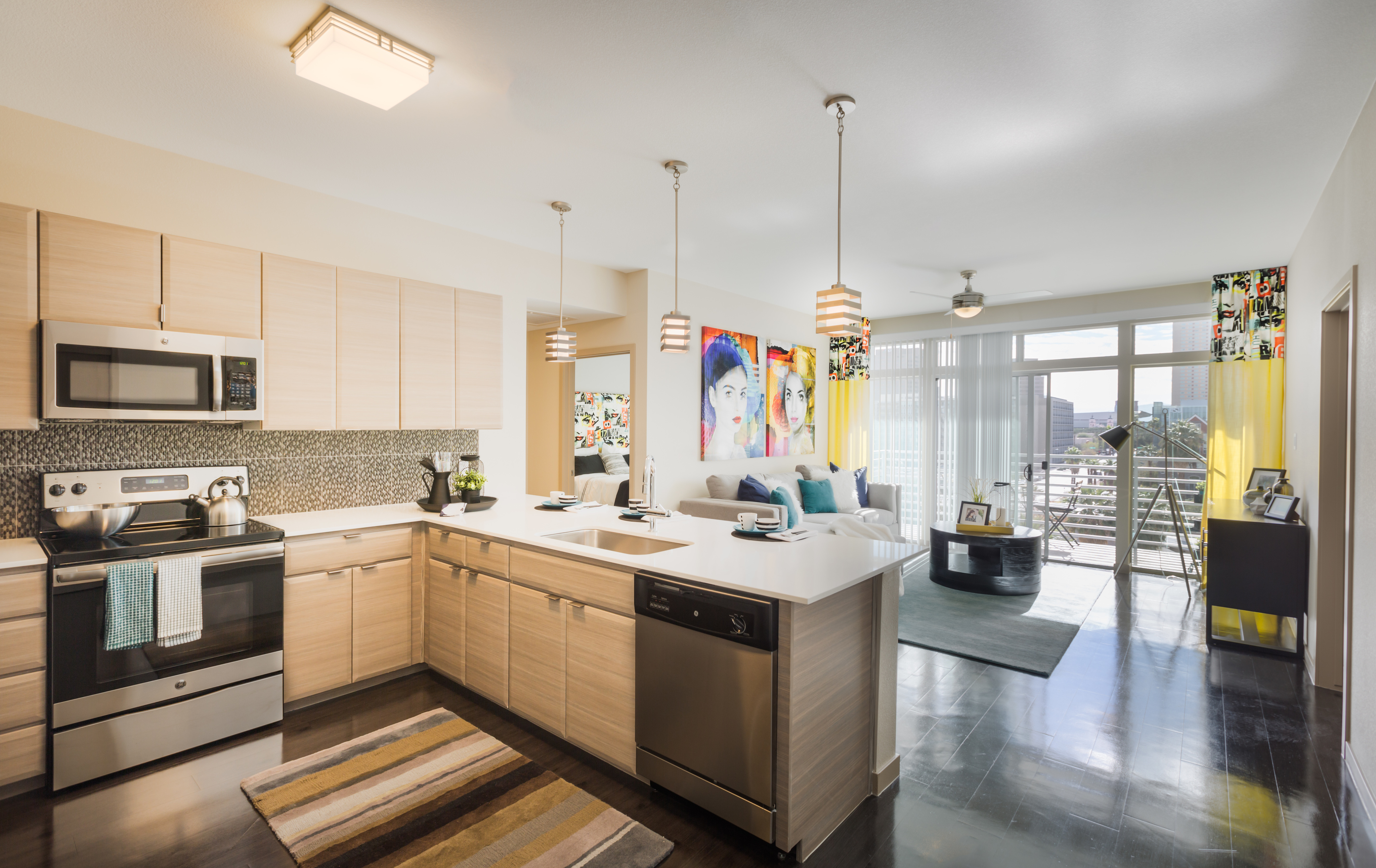 Modern open-plan kitchen and living space featuring sleek cabinetry, stainless steel appliances, vibrant artwork, and a sunlit balcony view.
