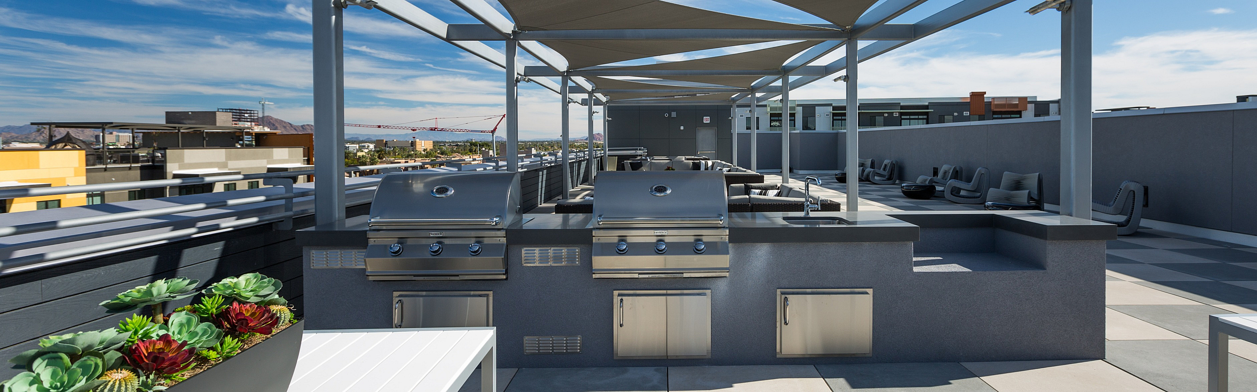 Contemporary rooftop patio with sleek outdoor grills, modern seating area, and pergola under a clear blue sky.