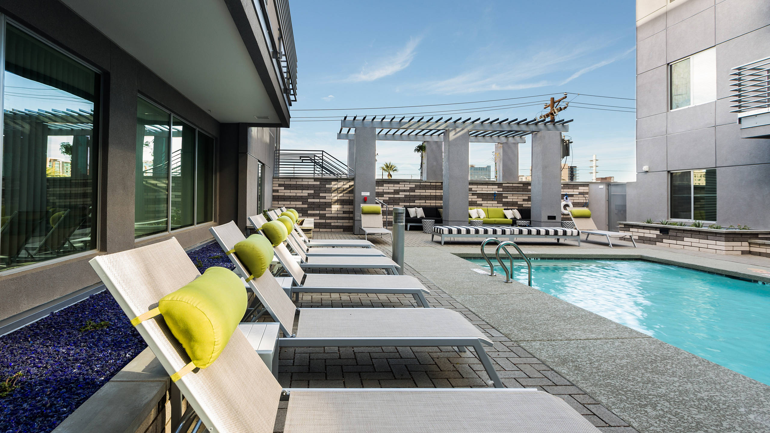 Modern apartment pool area featuring sleek lounge chairs with vibrant green cushions and a stylish pergola, perfect for relaxation and leisure.