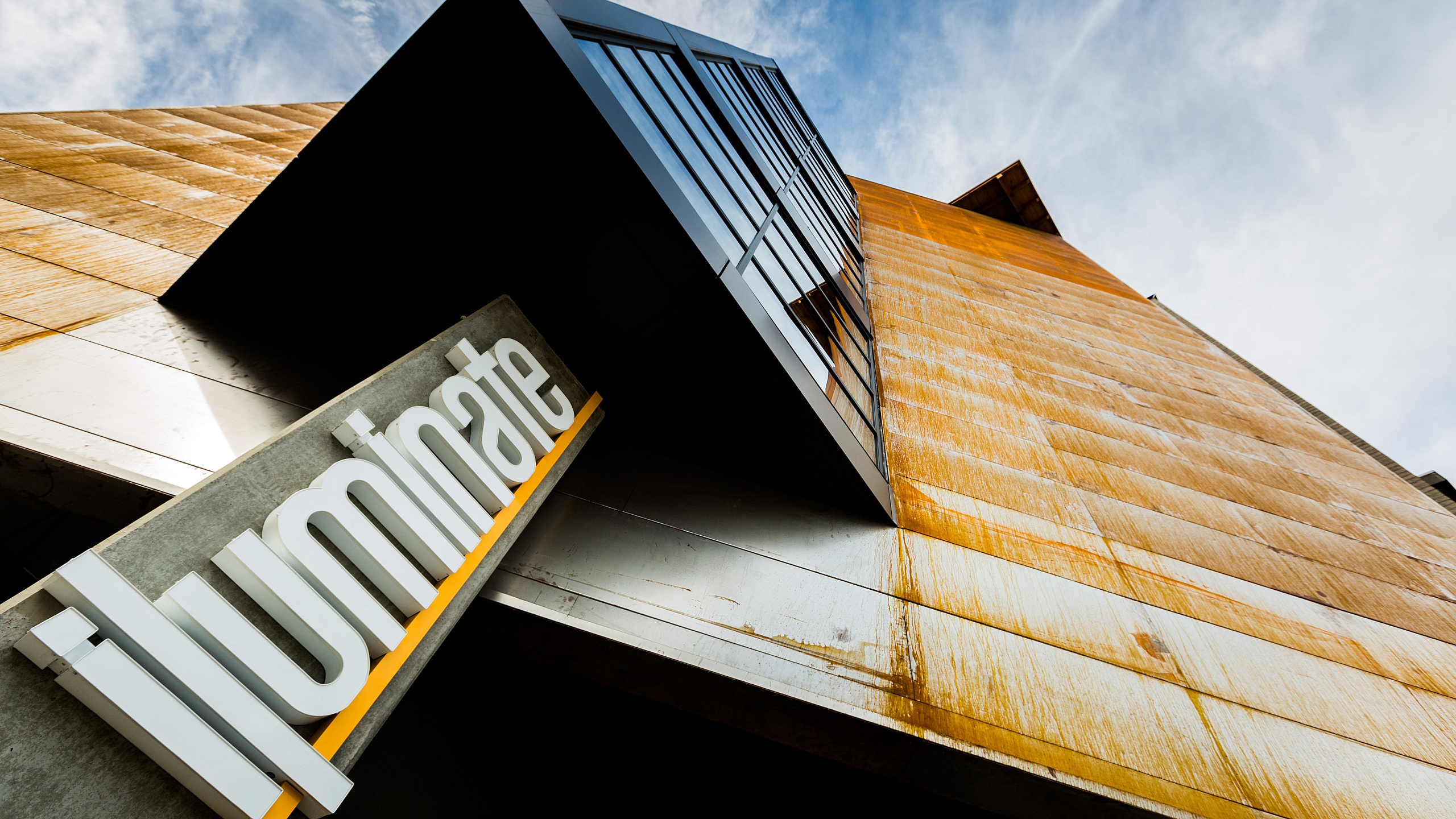 Dynamic perspective of a modern building facade with bold geometric windows and rust-colored panels against a blue sky.