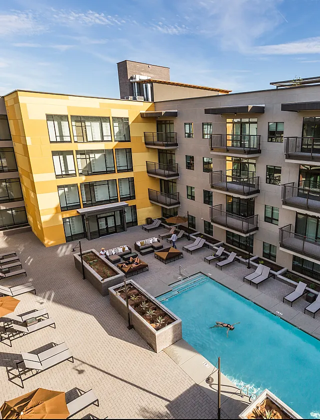 Modern apartment complex with vibrant yellow accents, featuring balconies overlooking a pristine outdoor pool and a sun-soaked lounge area.