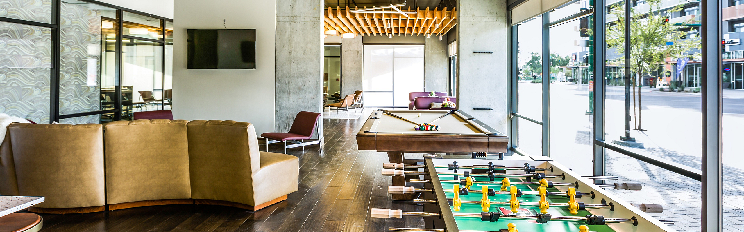 Modern apartment recreation room with sleek pool table, foosball, cozy seating, and floor-to-ceiling windows offering vibrant city views.