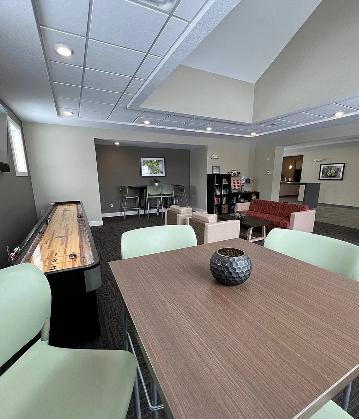 Modern recreational room with a shuffleboard table, stylish green chairs, and comfortable seating area, perfect for relaxation and entertainment.