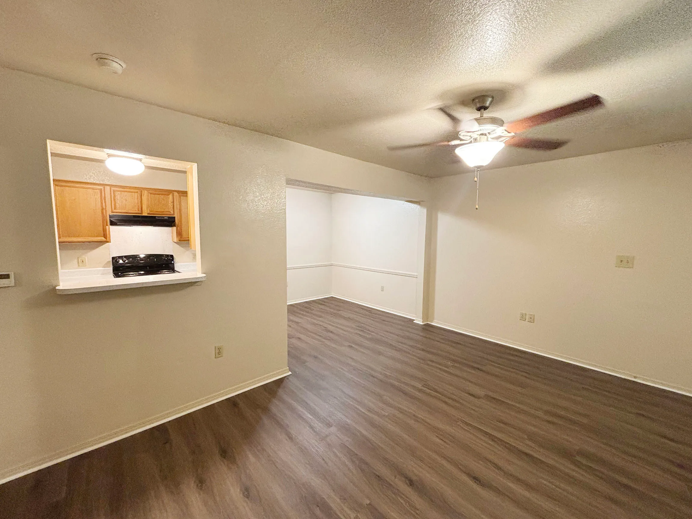Elegant open living space with wood flooring, ceiling fan, and kitchen pass-through window showcasing warm cabinetry and modern lighting.
